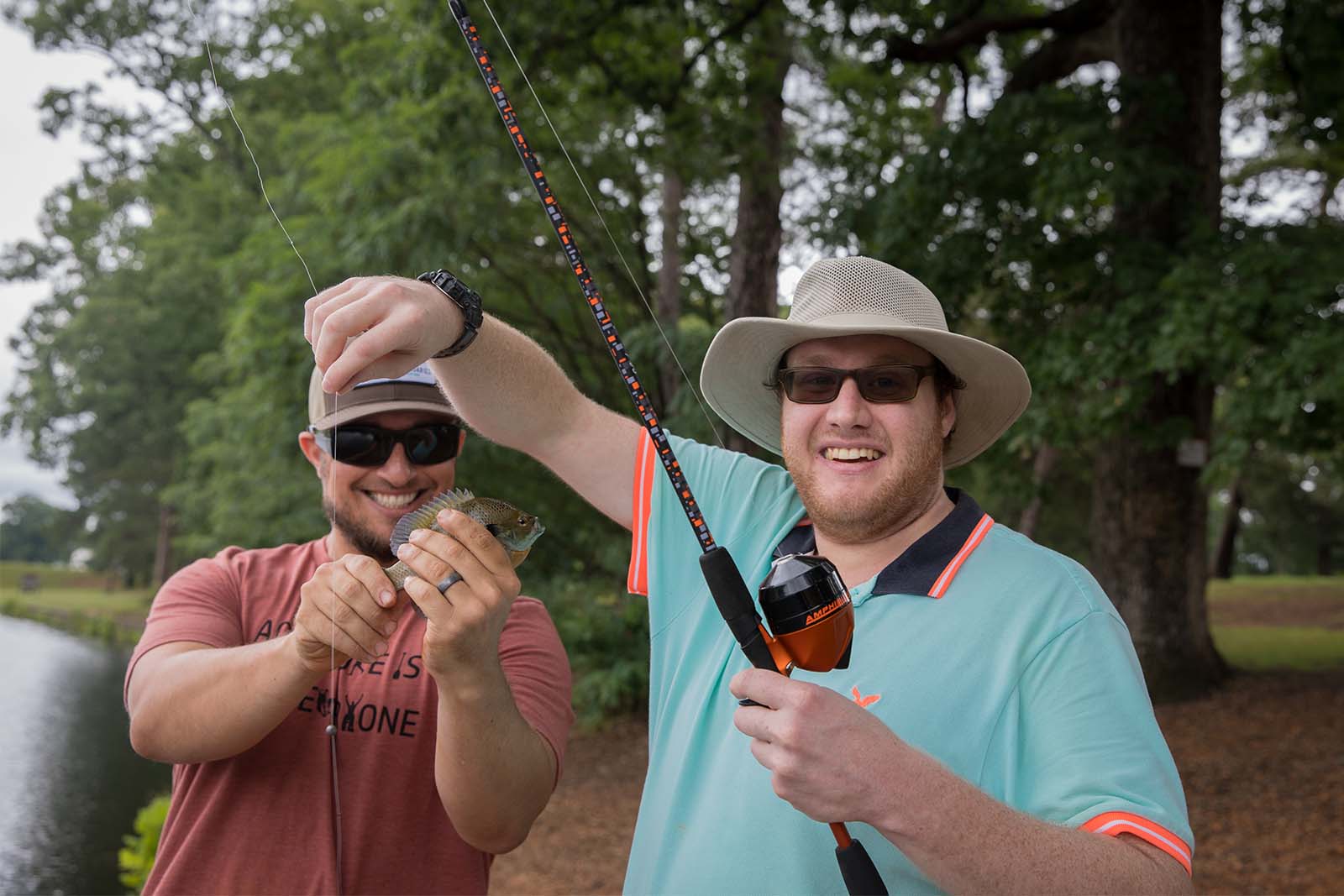 رجلان يقفان على شاطئ بحيرة. يمسك أحد الشخصين بصنارة صيد بينما يمسك الآخر بسمكة تم إزالتها من الصنارة