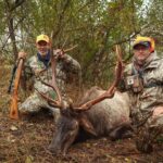 رجلان يقفان مع أيّل نافق، ممسكين برأسه من قرونه.