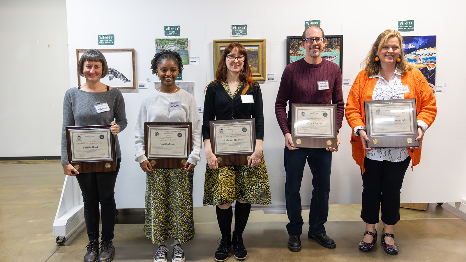 صورة لخمسة أشخاص يقفون حاملين شهادات أمام حائط عليه أعمال فنية.