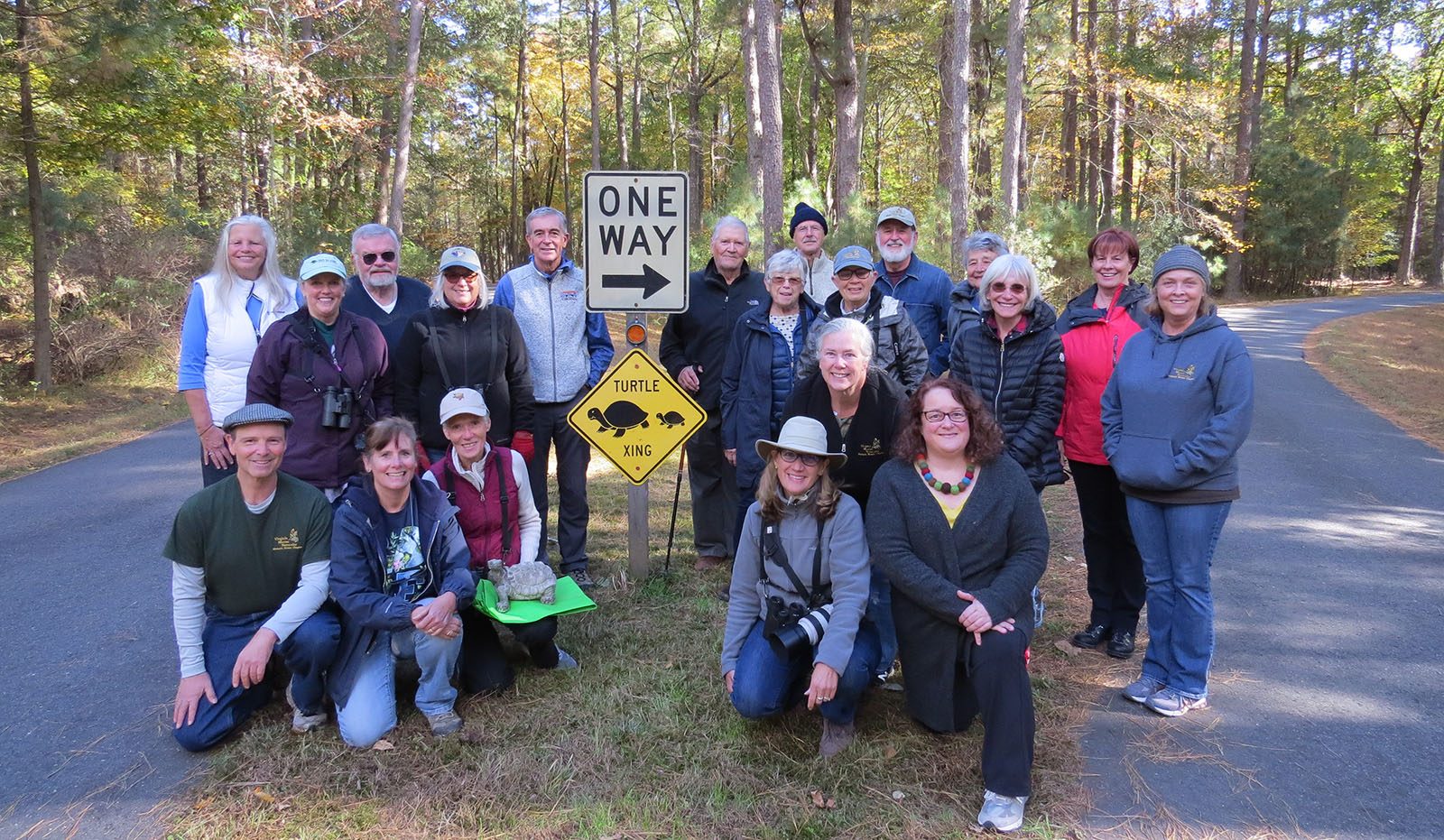 تجمعت مجموعة من 20 شخص حول علامة عبور السلاحف في منتصف طريق مرصوف صغير، وكان شخص واحد راكعًا في المقدمة مع تمثال سلحفاة.
