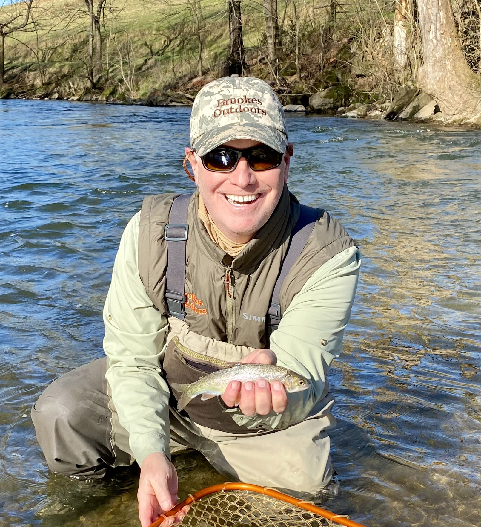 صورة لرجل راكع على ركبتيه في النهر، وهو يحمل سمكة تراوت قوس قزح صغيرة من شبكة.