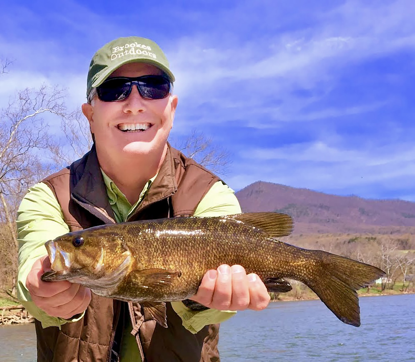 رجل مبتسم يحمل سمكة باس كبيرة من سمك القاروس الصغير خارج الماء بكلتا يديه والنهر في الخلفية.