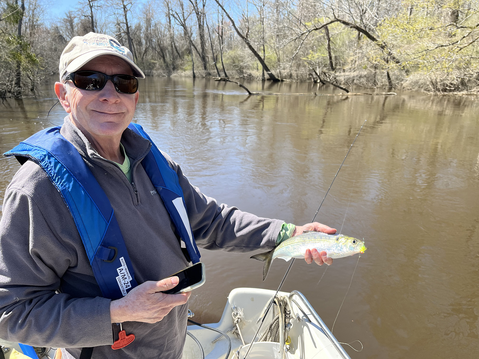 رجل يرتدي سترة نجاة ويقف على قارب في نهر ويحمل سمكة شاد لامعة.