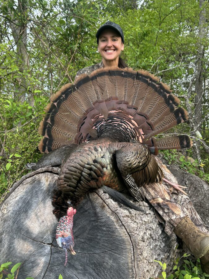 صورة لصياد أنثى تبتسم خلف ذكر ديك رومي كبير مستلقٍ على جذع شجرة.