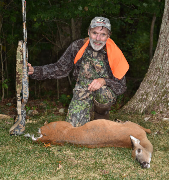 صورة لرجل يرتدي ملابس مموهة وبرتقالية ويحمل بندقية ويركع خلف ظبية بيضاء الذيل على الأرض.