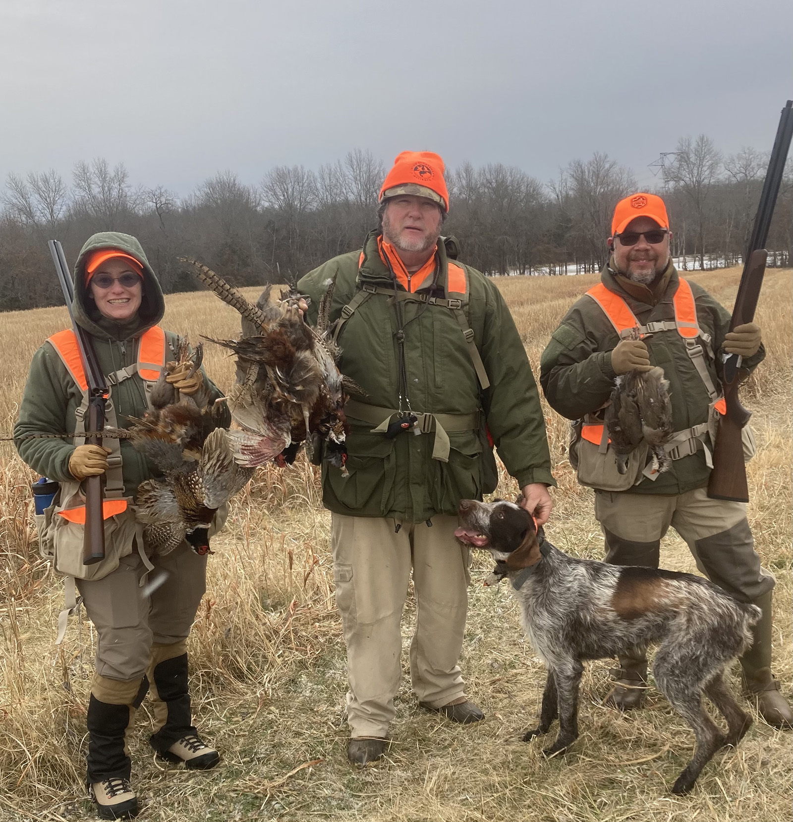 A photo of a woman and two men standing in hunting attire and carrying guns and harvested birds.