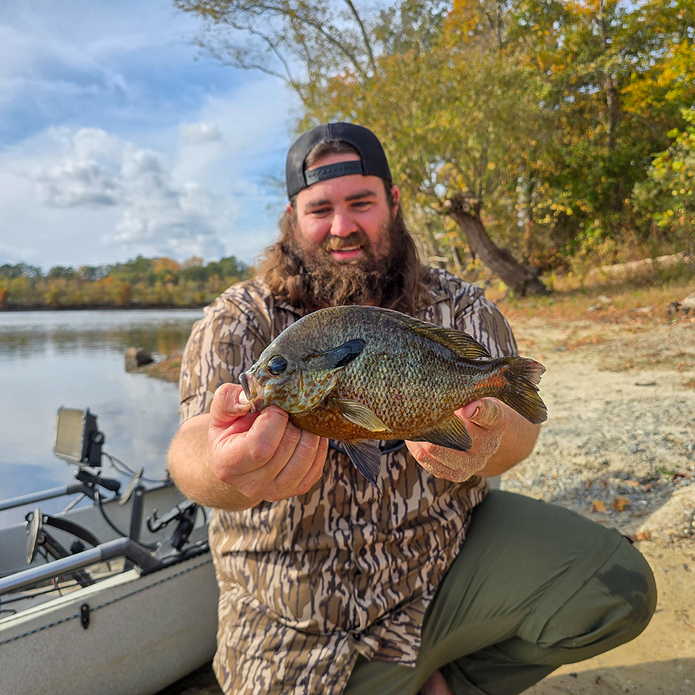 صورة لرجل يجثو على ركبتيه أمام زورق على الشاطئ، وهو يحمل سمكة شمس حمراء الصدر كبيرة الحجم ويبتسم.