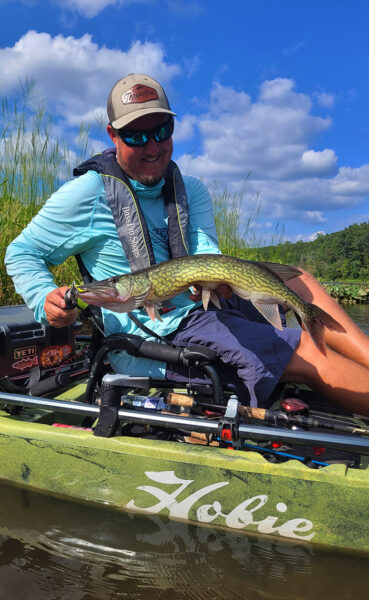 رجل في زورق صيد في قارب كياك يجلس على السطح ويحمل سمكة بيكريل خضراء.